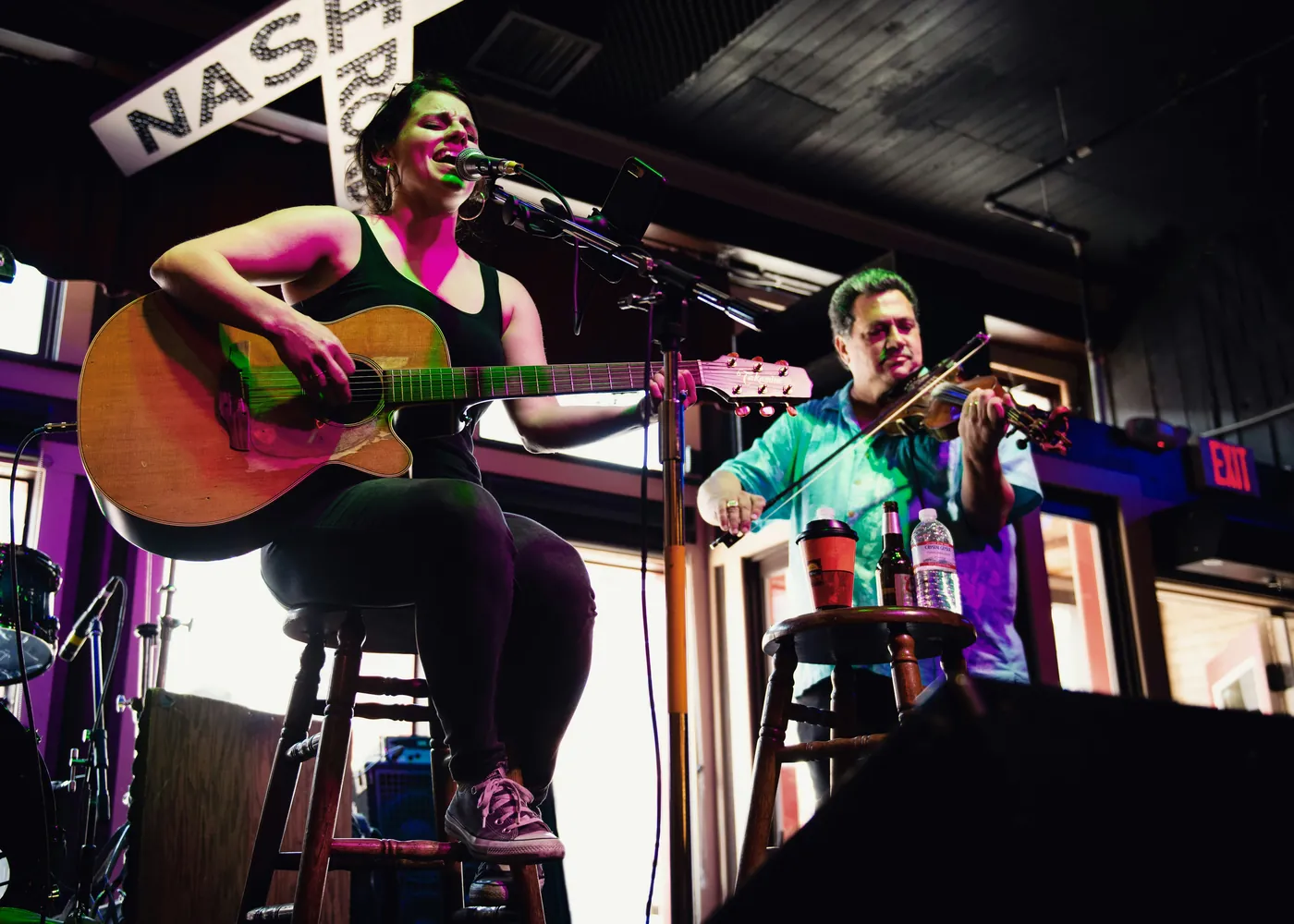 A singer with an acoustic guitar and a fiddle player performing on stools at a small venue