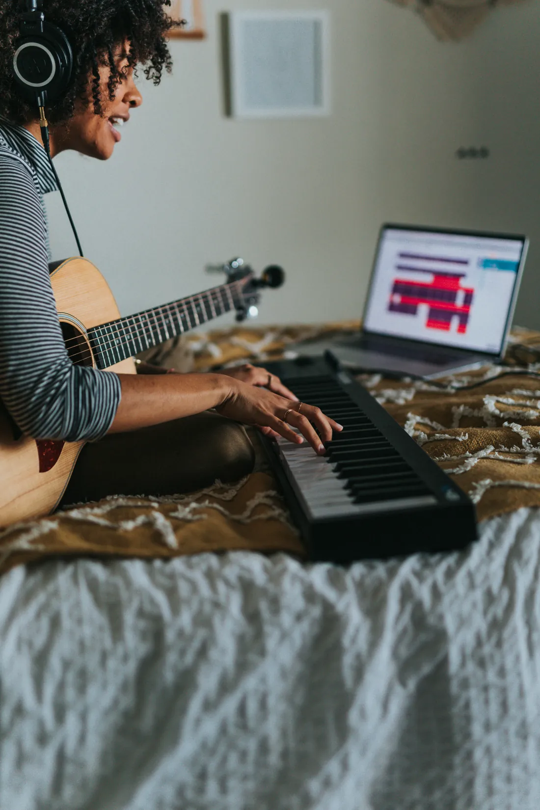 A musician with headphones writing a song at home, playing a keyboard with an acoustic guitar and a laptop.